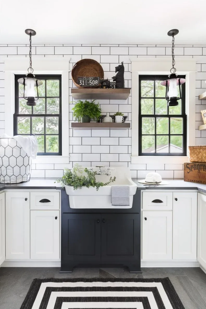 White apron-front utility sink below rectangular tile wall between black-framed windows, with dark sink cabinet and hanging lanterns