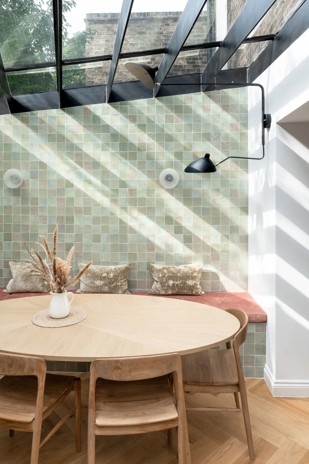 A pale rounded dining table and curved wood chairs stand beside a cushioned bench backed by pale green grid tiles beneath sloping glazing and projecting dark lights