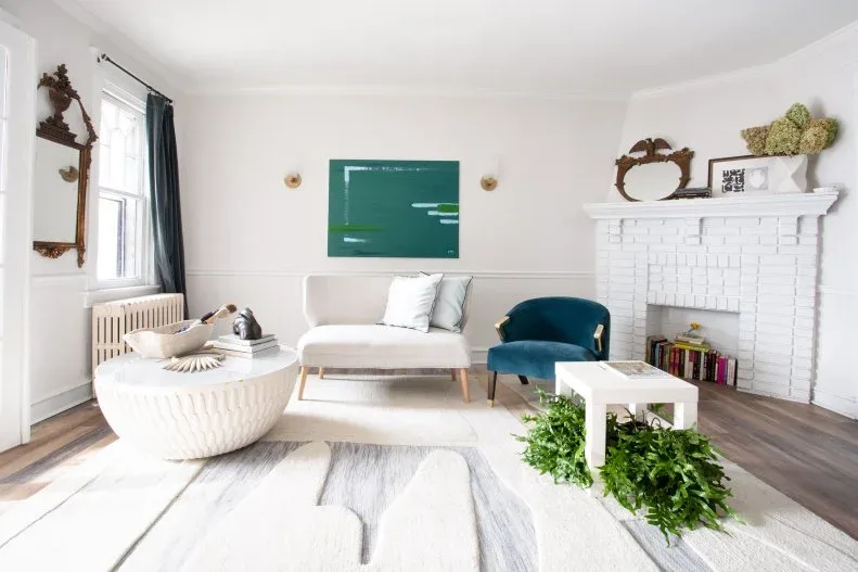 White living room with round ribbed table, pale curved settee, teal armchair, green artwork, white brick fireplace and dark decorative mirror.