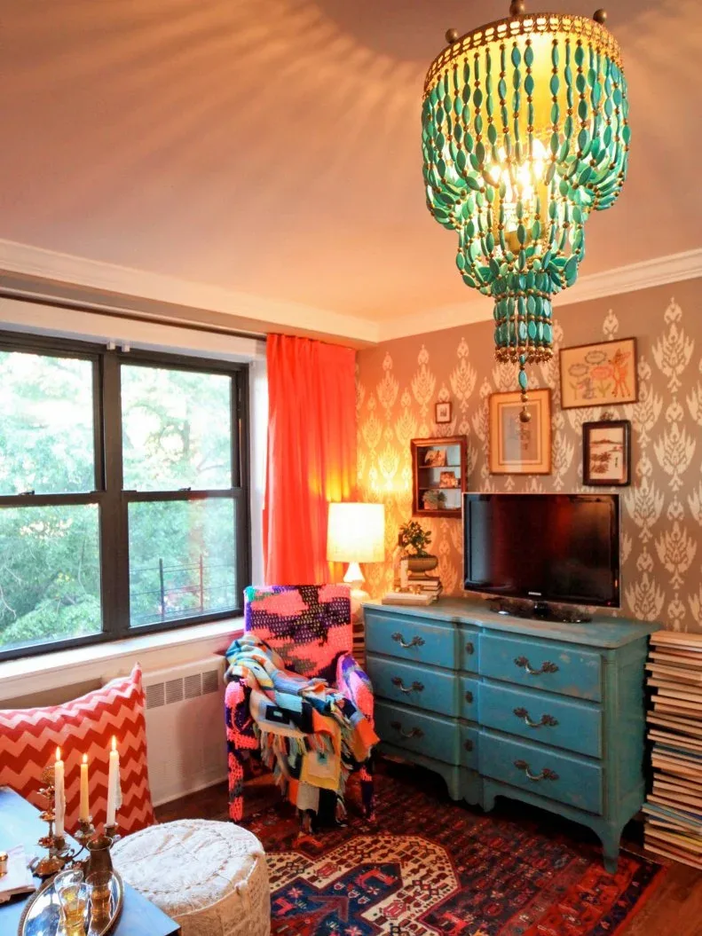 Living room with turquoise drawer cabinet and beaded pendant, orange curtain, patterned brown wall, colorful armchair, television and layered patterned rug.