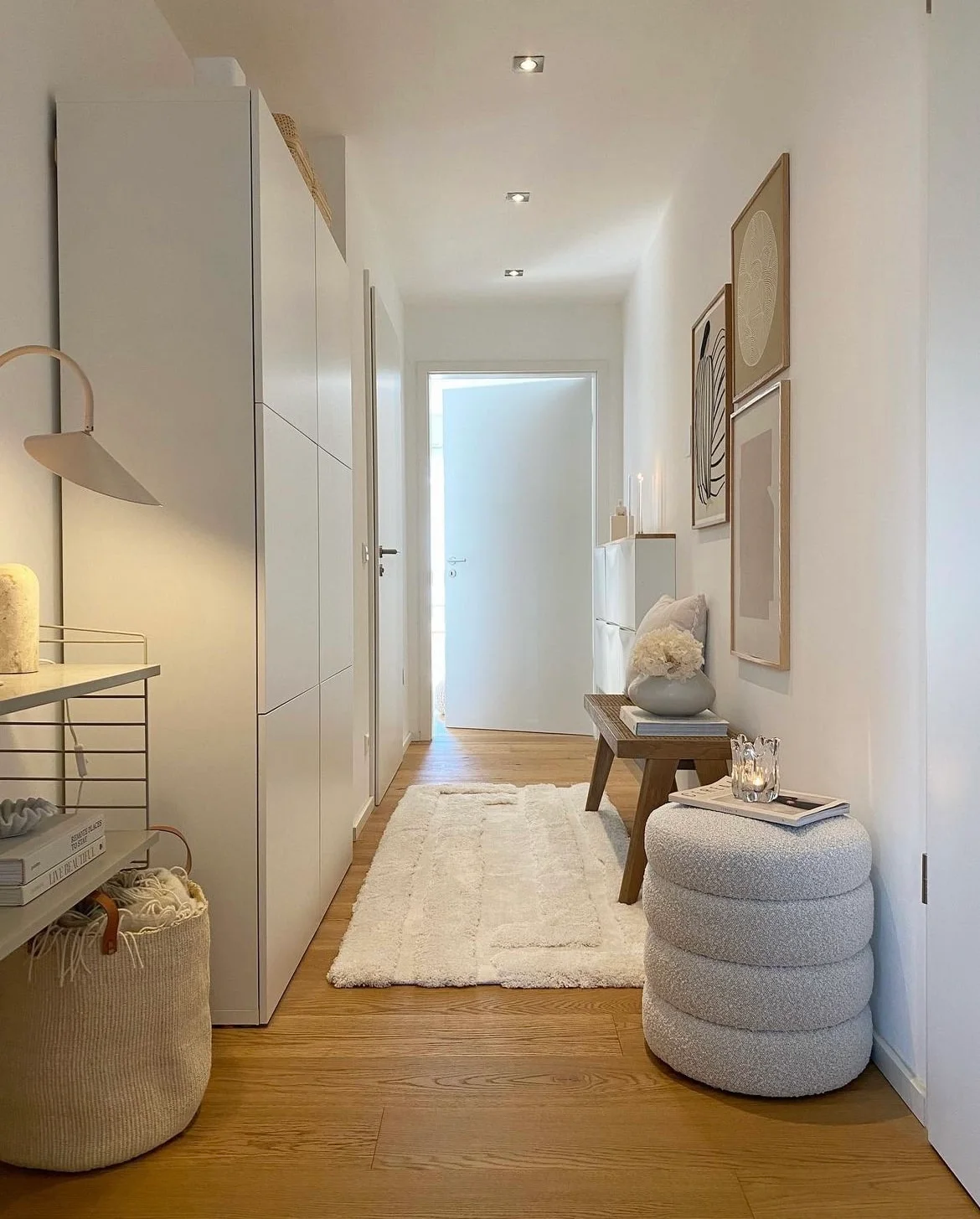 Tall pale cabinets line a narrow wooden-floored hallway opposite a small wooden bench, rounded gray stool and framed pictures