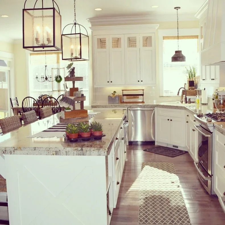 White kitchen with large pale island, dark framed lantern pendants, wood floor and small plants beside woven-backed stools