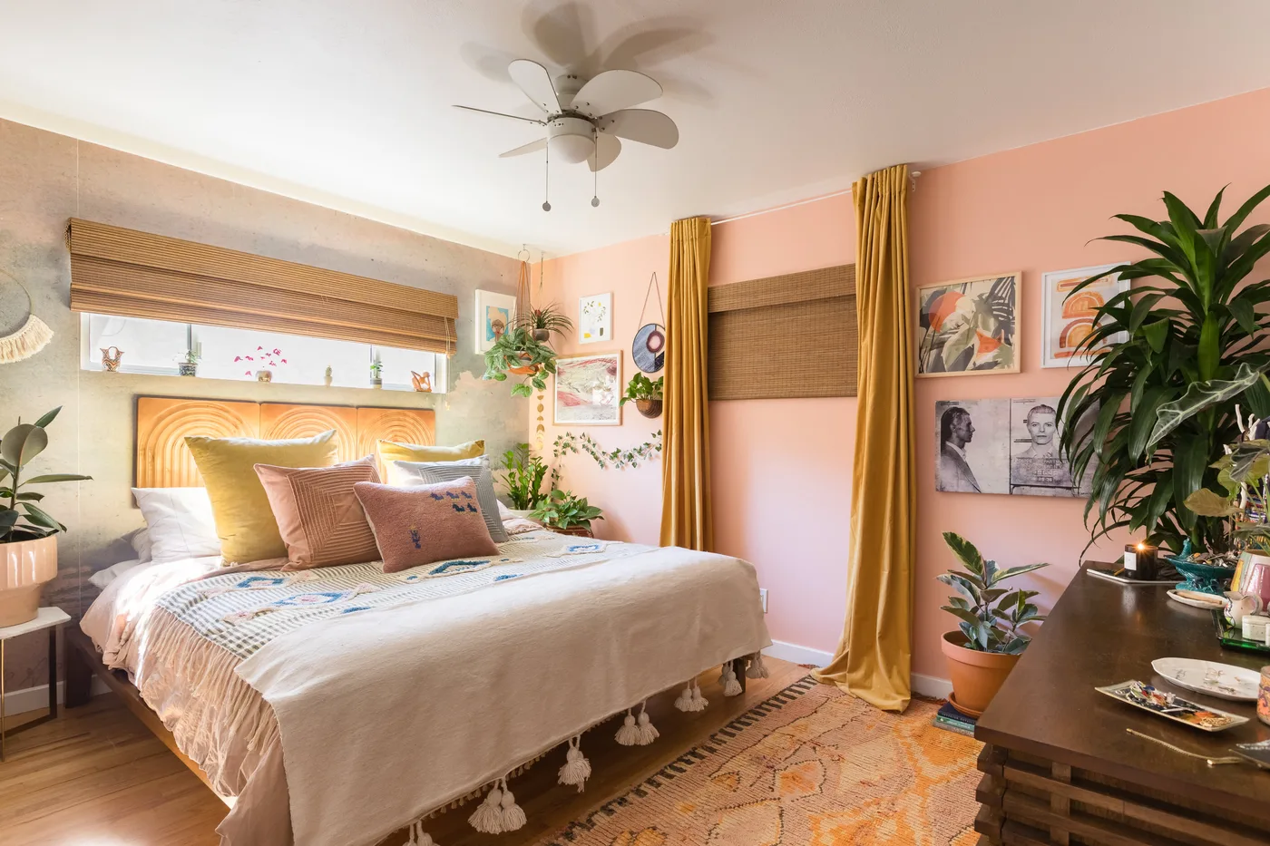 Bedroom with pink wall, mustard curtains, wooden headboard, pale layered bedding, several potted and trailing plants, and dark wooden dresser.