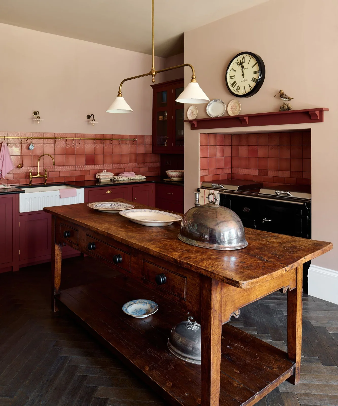 Deep red cabinets and reddish wall tiles surround a wooden worktable island beside a white apron-front sink and dark cooker