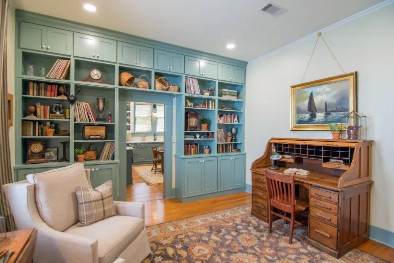 Country study with wood rolltop desk, blue-green fitted bookcases, pale armchair and patterned rug