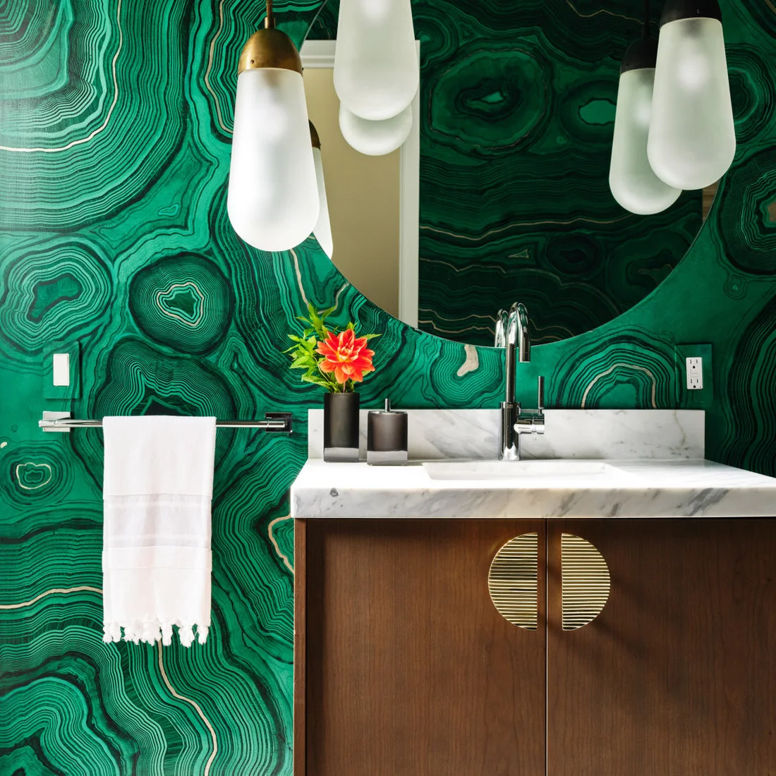 Green contour-like wall pattern surrounds a large round powder-room mirror and pale hanging lights above a wooden vanity with a circular gold-colored handle