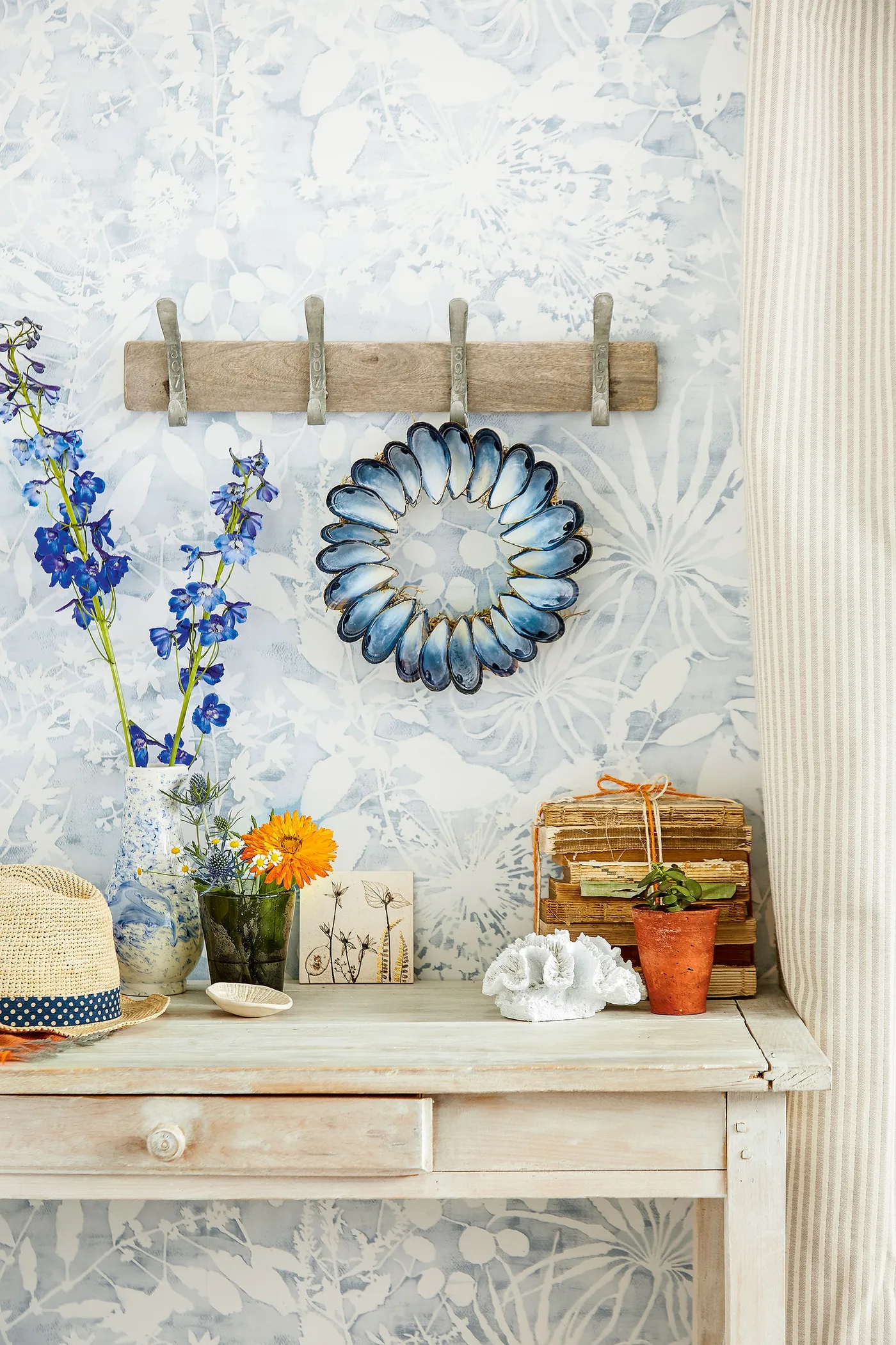 Country entry detail with pale leafy wallpaper, a wooden peg rail, blue-edged round mirror and a pale console holding flowers and small containers.