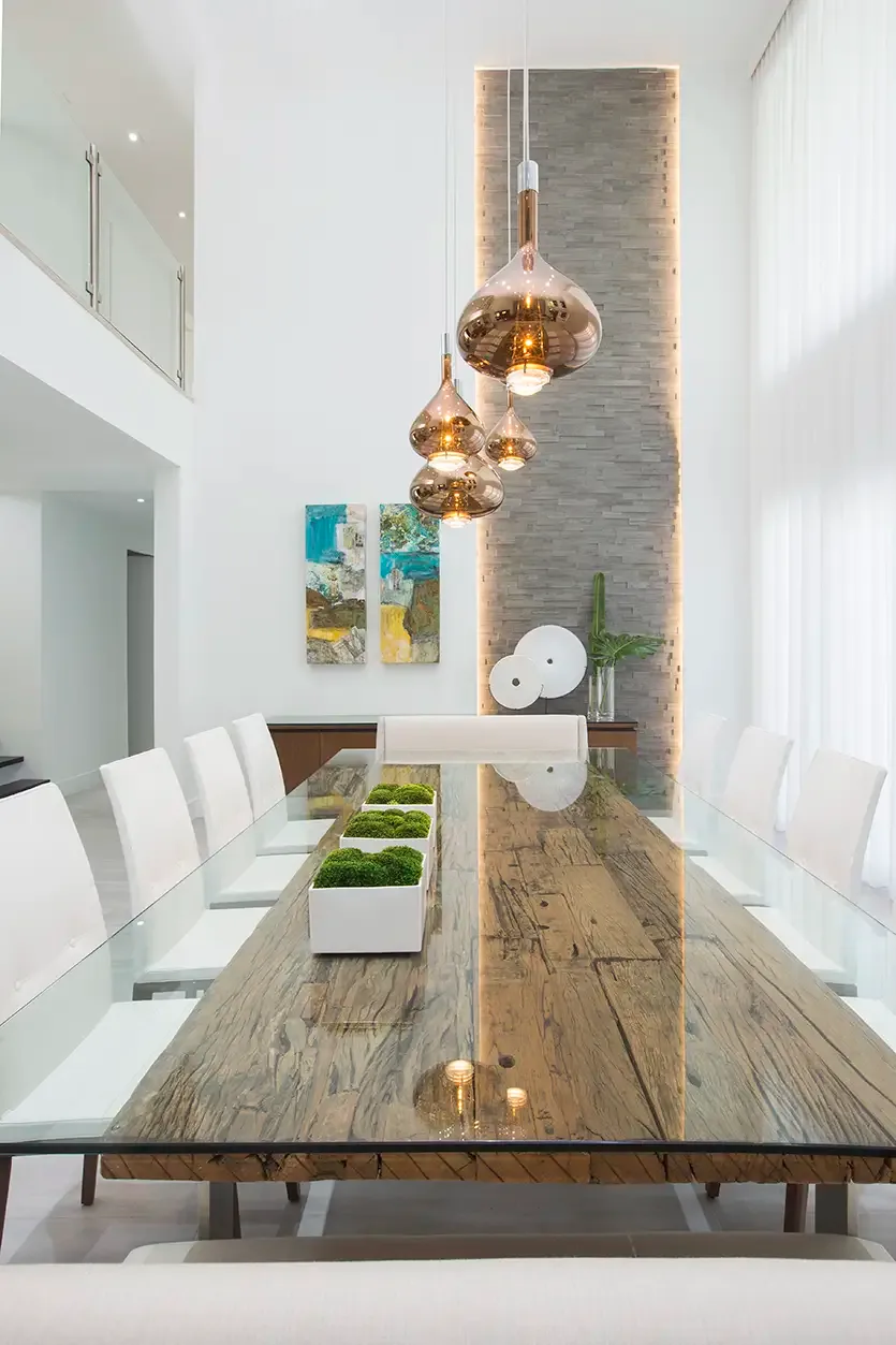 Long clear glass tabletop above a broad textured wood-toned center with white chairs and warm metallic pendants
