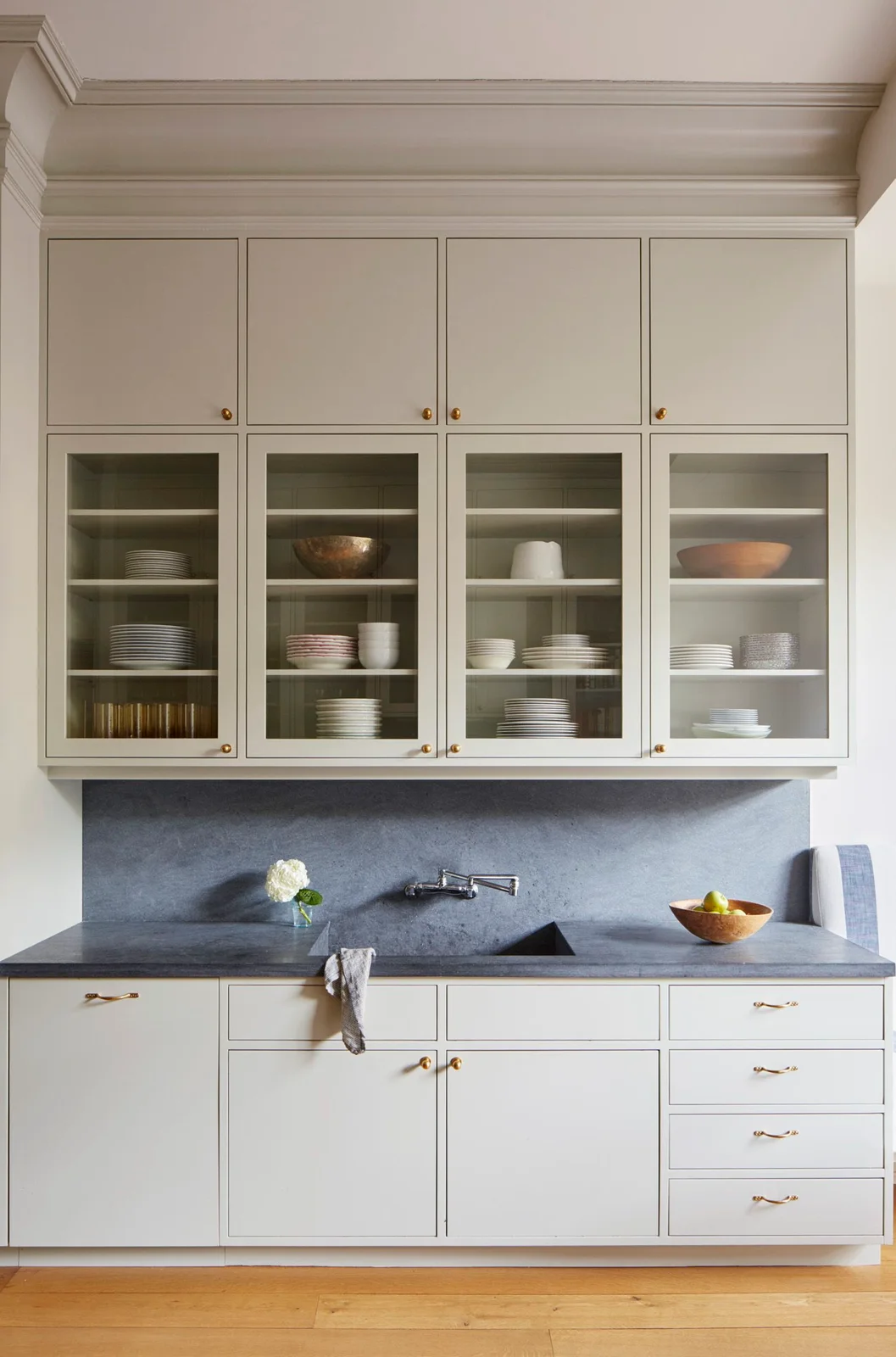 Light beige kitchen cabinets with glass upper doors sit above a dark counter and backsplash beside a warm wooden floor