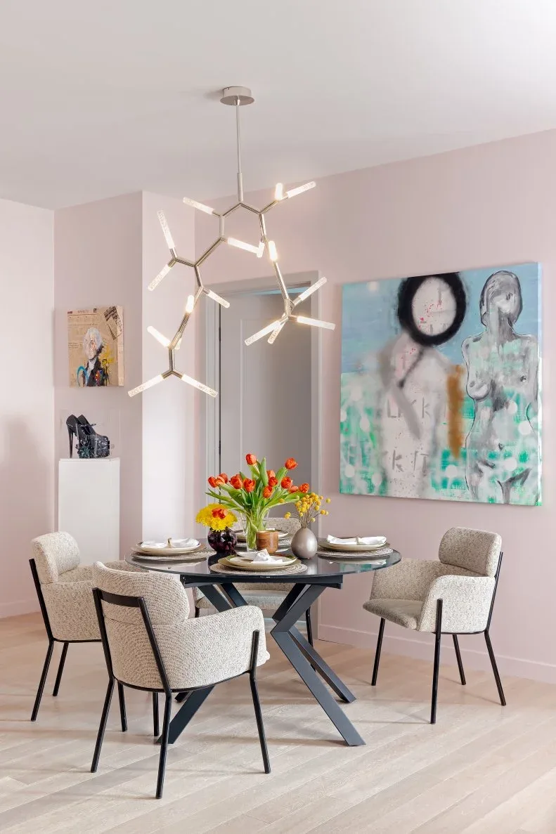 Round glass dining table on a dark crossed base with gray upholstered chairs in a pink room.