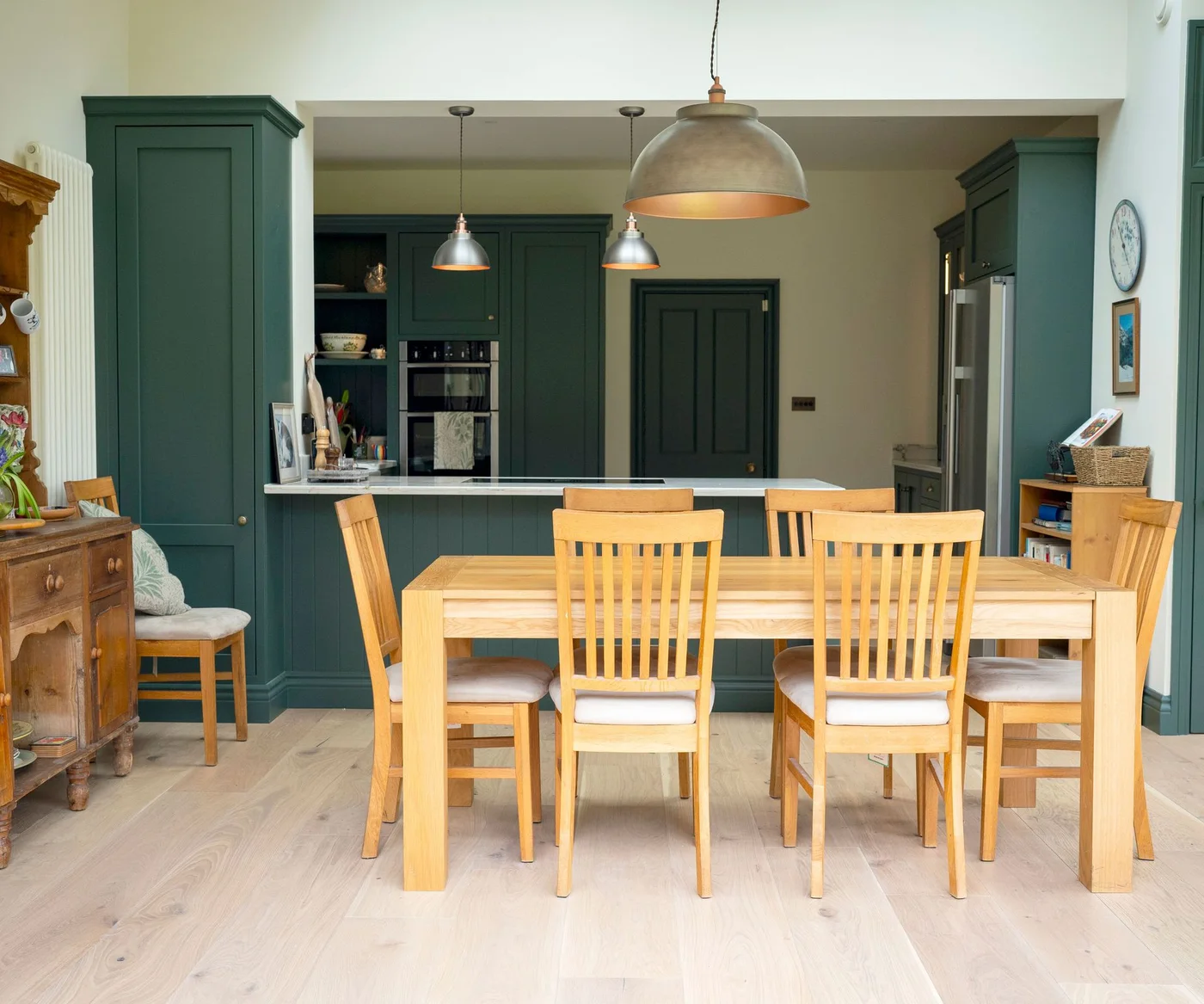 A wooden dining table with matching chairs stands in front of dark green cabinets and a pale-topped peninsula