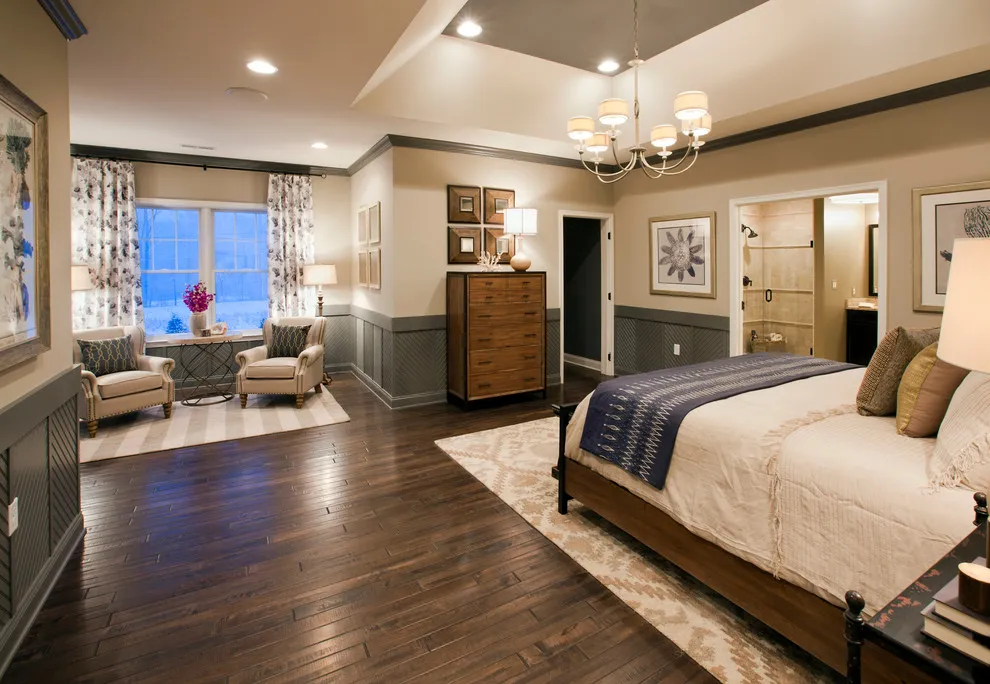 A beige bed sits above a dark wood floor beside pale chairs and framed art