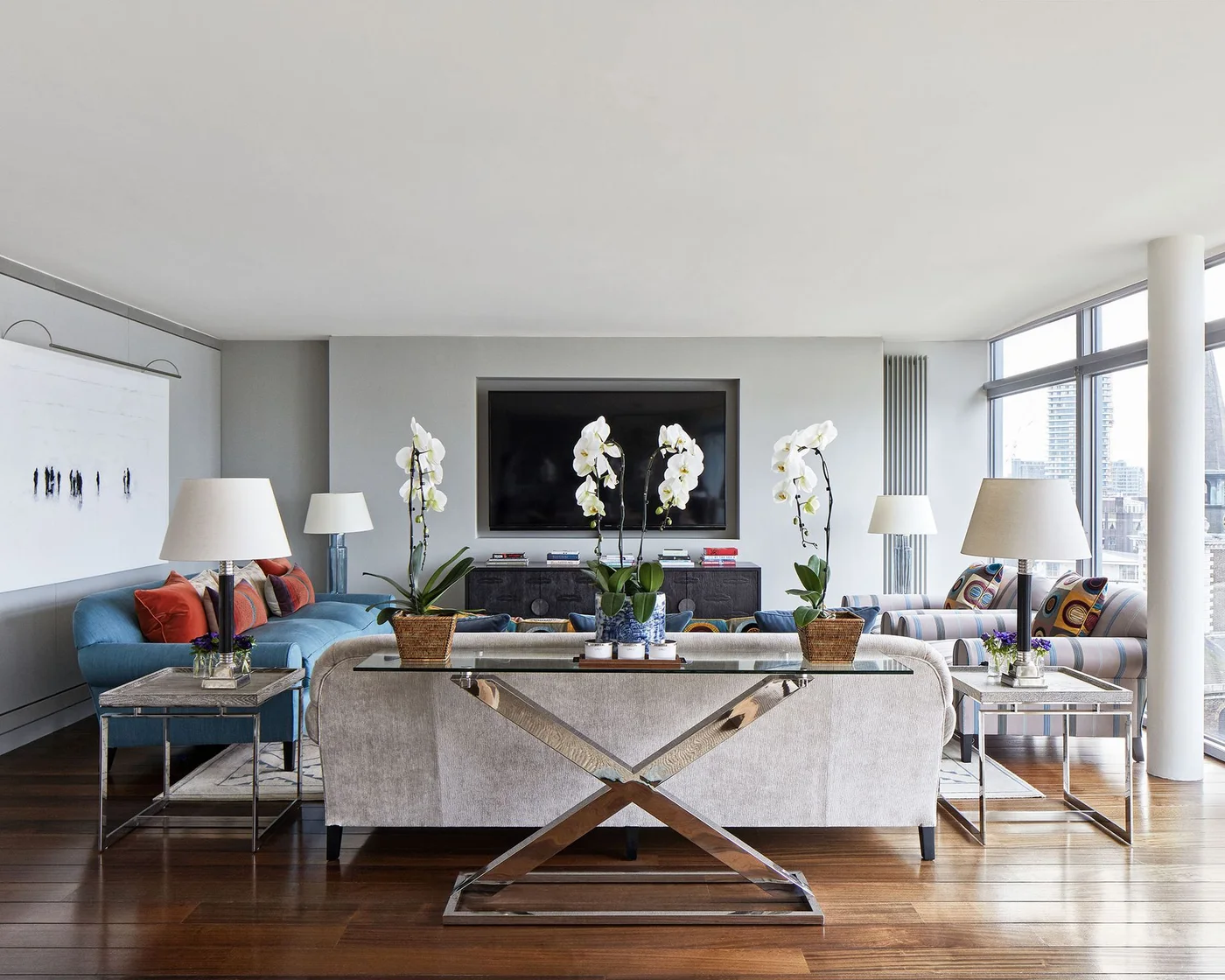 A wall-mounted television faces pale seating and a blue sofa behind a console with white orchids and matching lamps