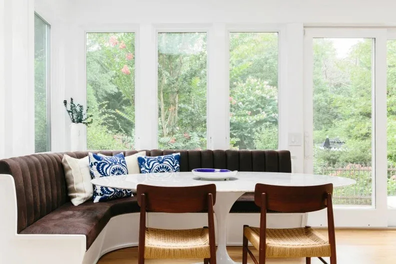 Dark brown corner banquette beneath tall windows, with a white oval table, blue-and-white pillows, and wooden chairs with woven seats.