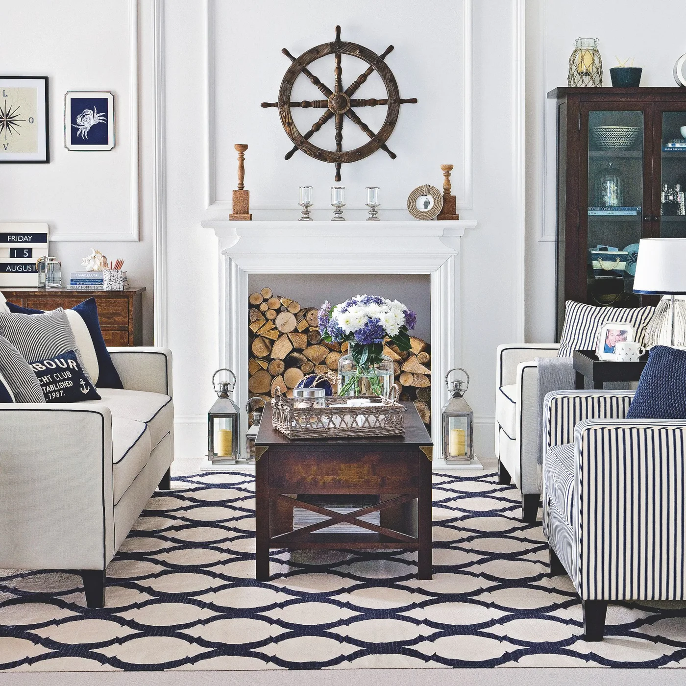 A ship wheel hangs above a pale fireplace between blue-and-white seating, dark cabinets and a wooden coffee table