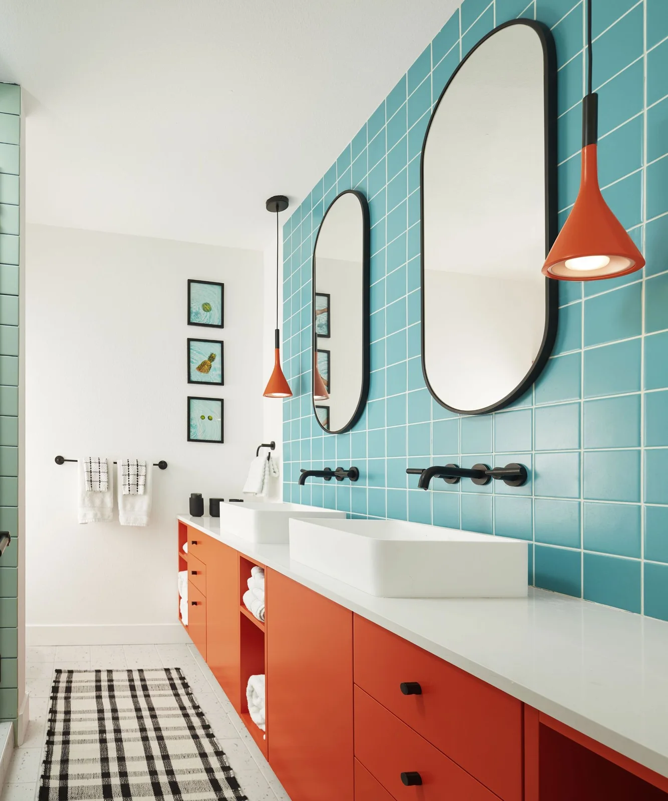 Blue square tiles form a backdrop for oval mirrors and orange pendant lights above orange cabinets and white vessel basins