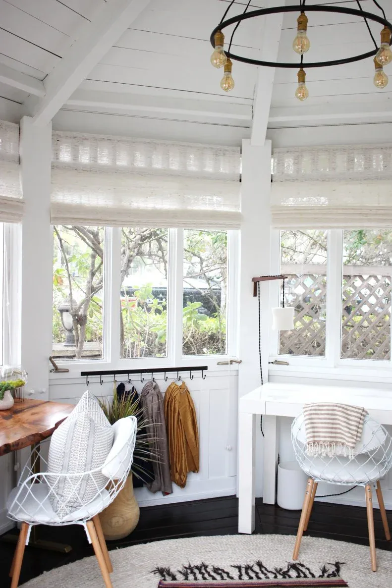 Bright garden workspace with separate white and wooden desks, open white chairs, individual lamps, woven shades, wall hooks and pale layered rugs.