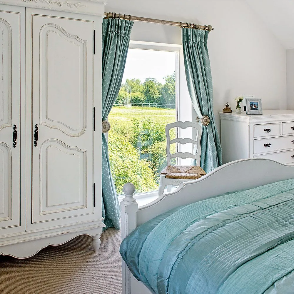 Pale bedroom with a decorative wardrobe, blue-green curtains and quilt, painted bed frame, white drawers and a wooden chair.