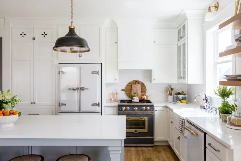 A dark range with warm-toned controls stands within pale framed cupboards beneath a plain pale hood beside an apron-front sink and open wood shelves