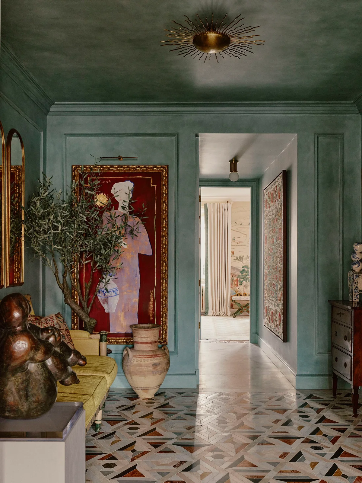 Green paneled walls surround a large gold-framed artwork, yellow seating, sculptures, a tall plant and a multicolored patterned entryway floor