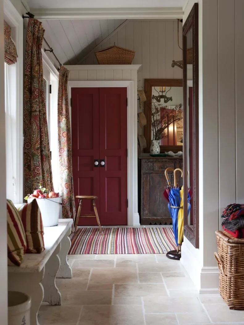 White board-lined mudroom with red cupboard, striped rug, curtained window, white benches and umbrella stand