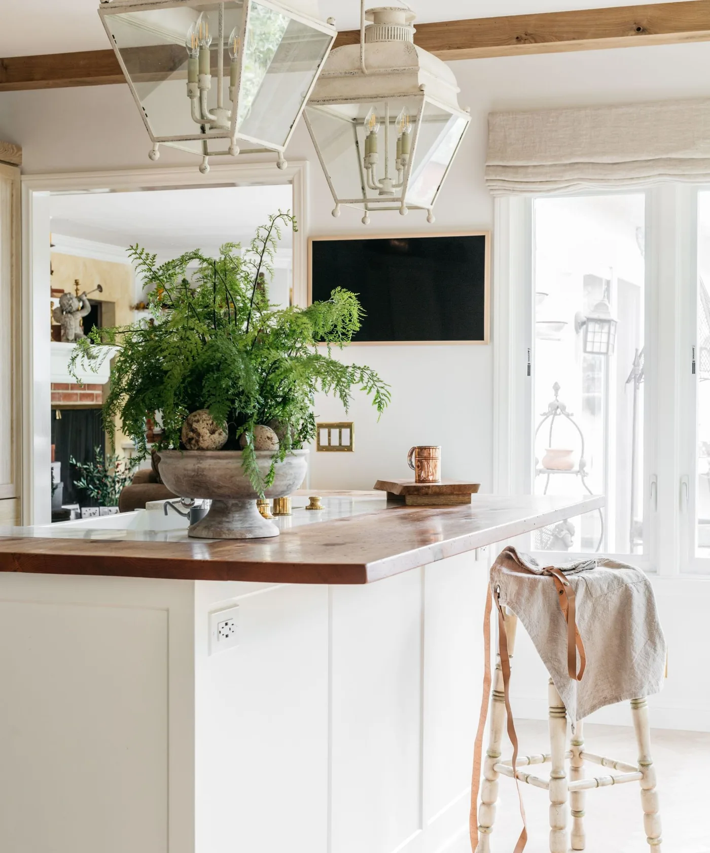 White farmhouse kitchen with a dark-topped island, woven stools, black lantern pendants and large fern.