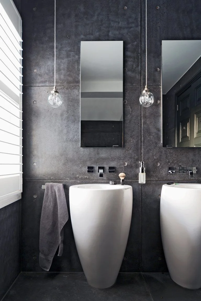 Two tall white pedestal basins stand against dark textured walls below narrow mirrors and hanging glass lights beside white window shutters