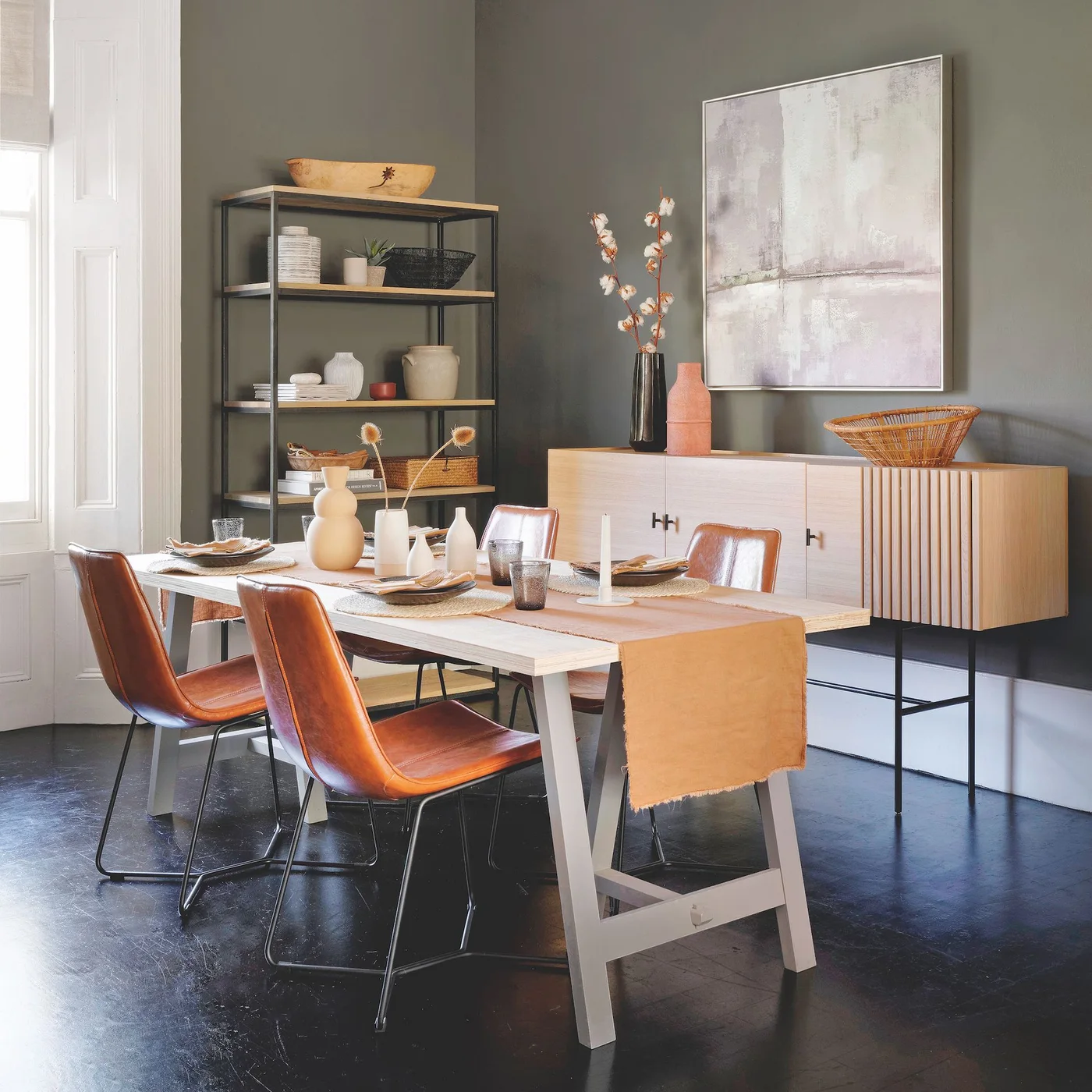 A pale wooden table and brown chairs sit against muted green walls beside a pale sideboard, open shelves and framed abstract artwork