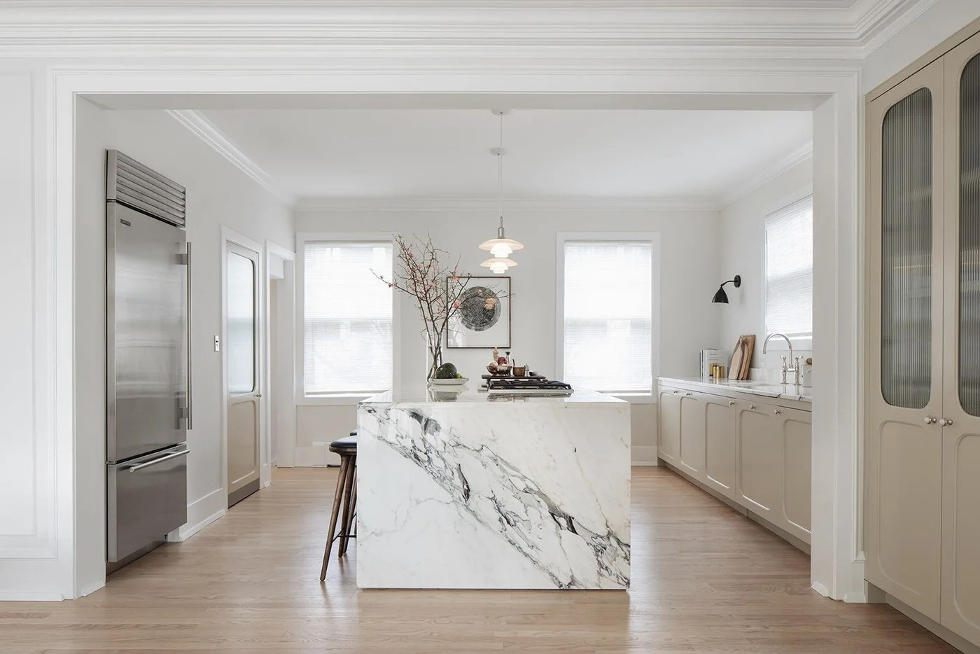 Pale beige perimeter cabinets surround a white island with dark veining beside a wooden floor and bright windows