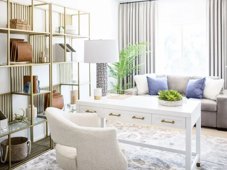 White office desk with gold-toned drawer pulls beside tall open metal shelving and a pale sofa.