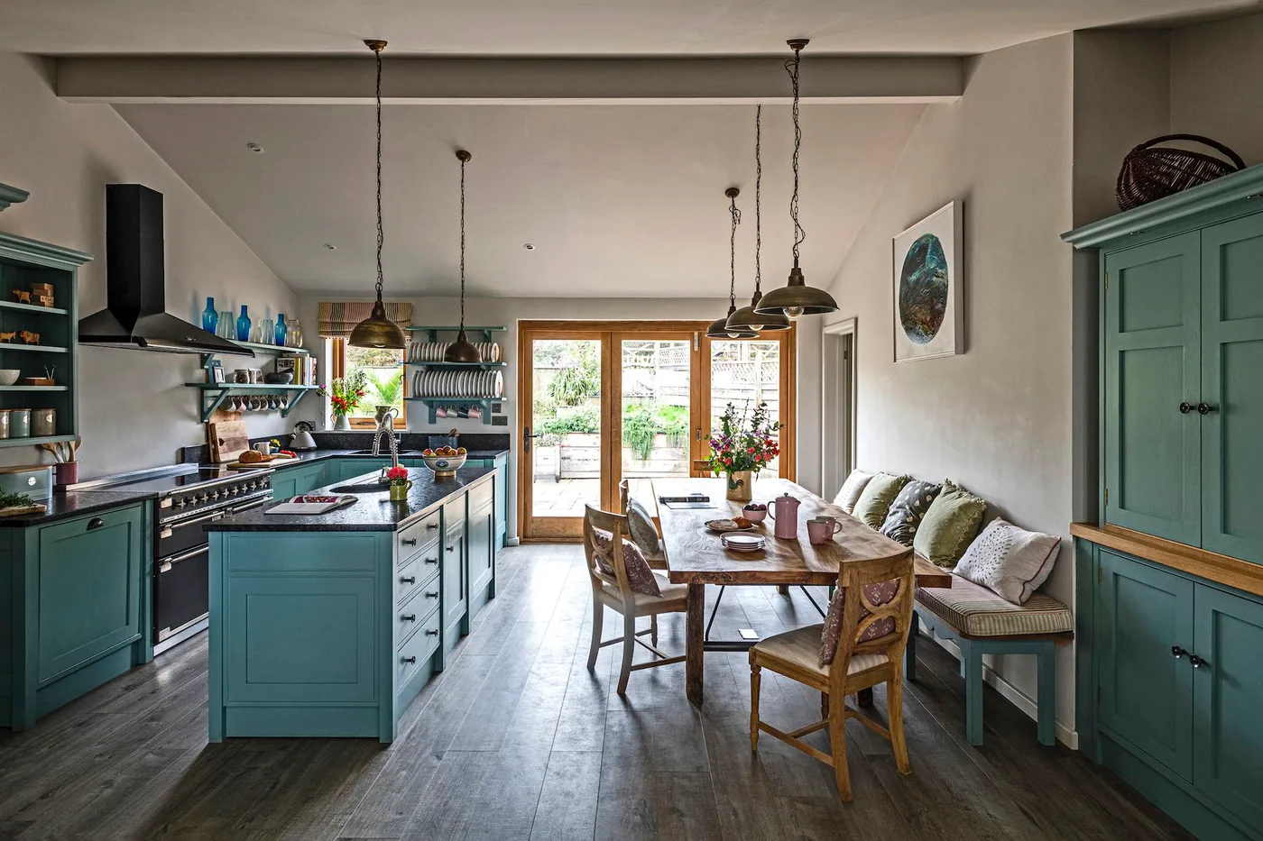Cottage kitchen with blue-green cabinets and island, wooden dining table, cushioned bench, dark floorboards and several warm metal pendant lights.