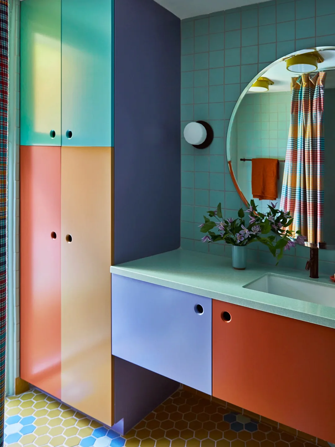 Turquoise, coral and purple bathroom cabinets surround a pale green counter beside a round mirror, grid tiles and striped shower curtain