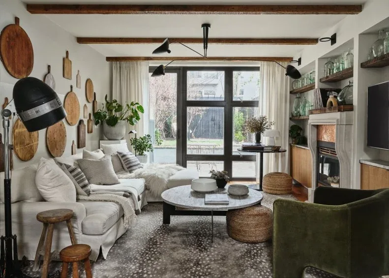 Country room with cutting-board wall display, pale sofa, round table, green chair and fireplace shelving beneath timber beams
