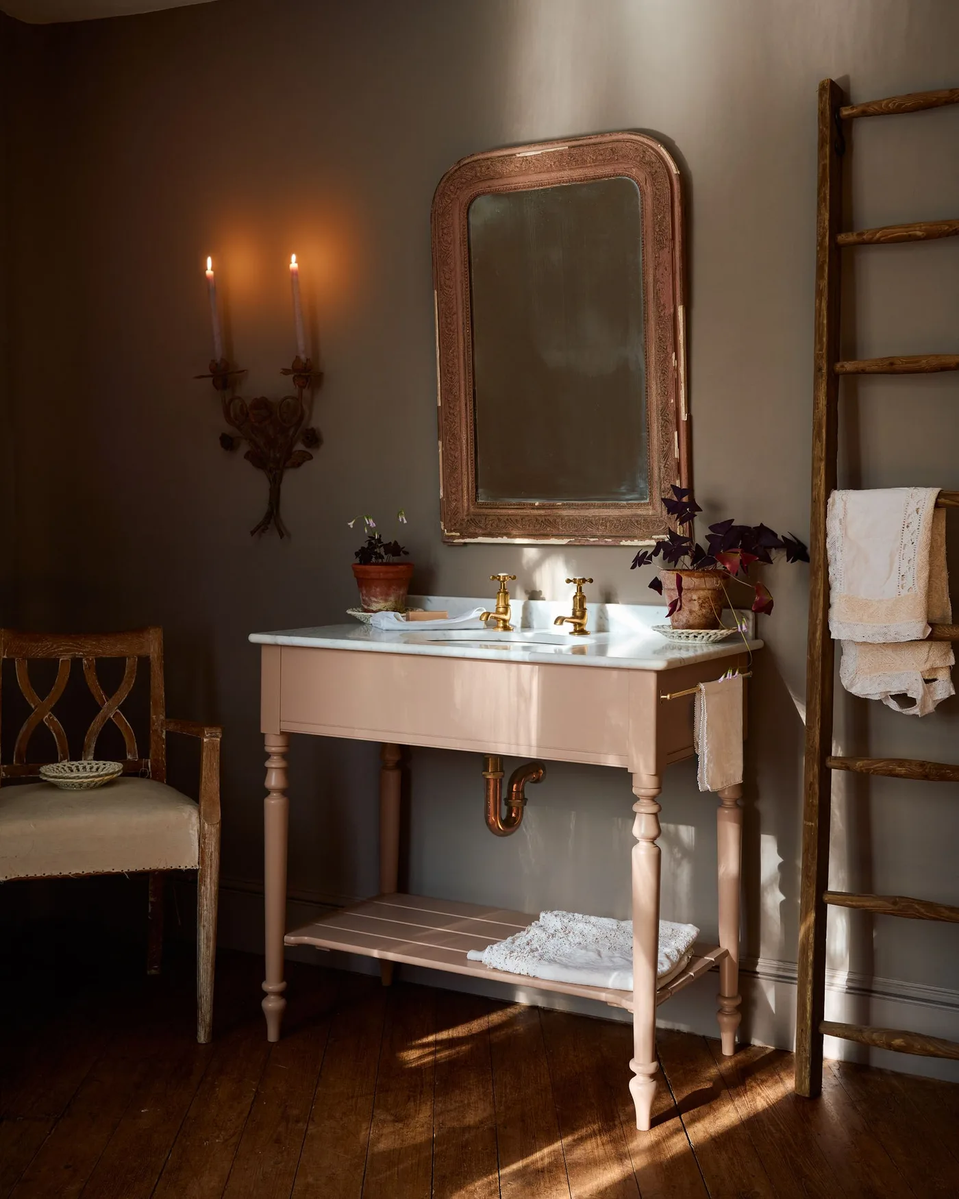 Muted brown bathroom wash area with pale turned-leg vanity, shaped mirror, wood ladder and chair