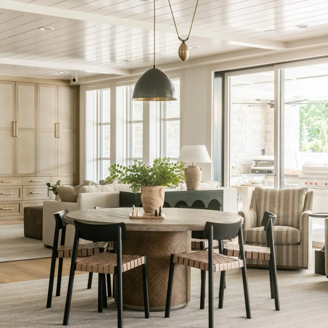 A pale round pedestal dining table and black chairs with woven seats sit beneath a gray dome pendant beside a light rug and pale living-room seating