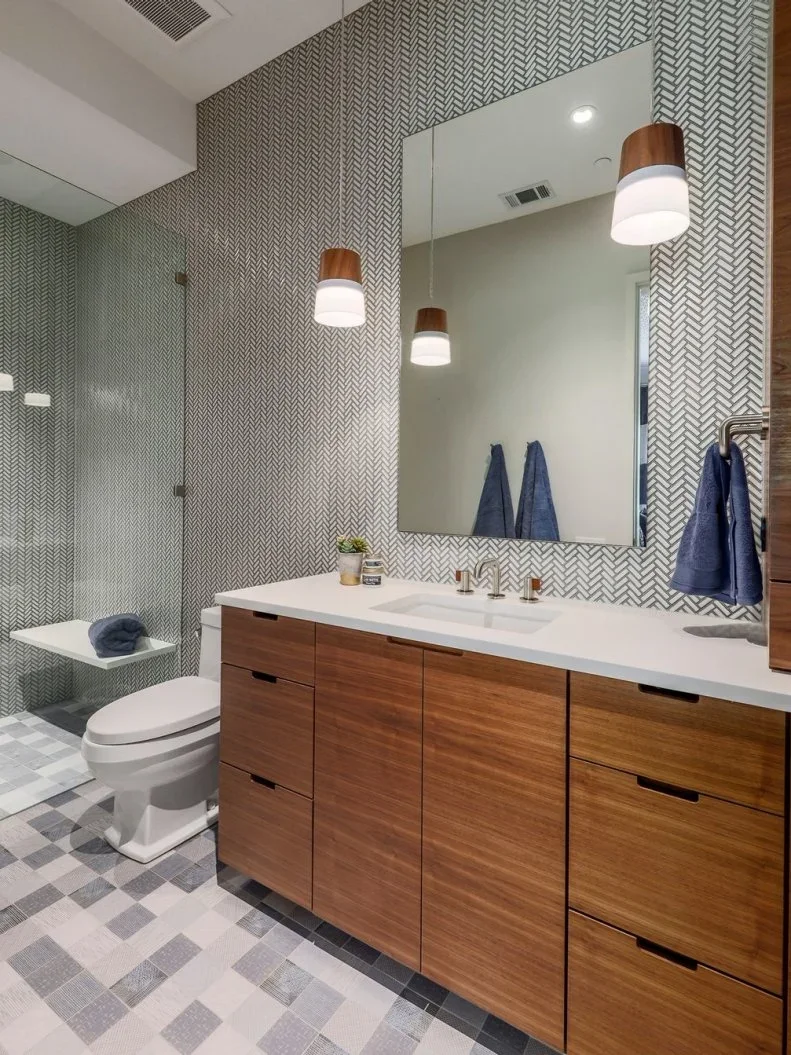 A wood drawer vanity stands beneath a broad mirror and small pendant lights against a herringbone-patterned wall beside a white toilet and clear shower screen