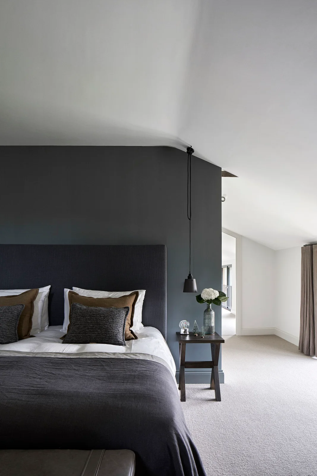 A dark headboard wall and gray bed covering sit beside white sloped ceilings, a wood bedside table and a hanging dark pendant