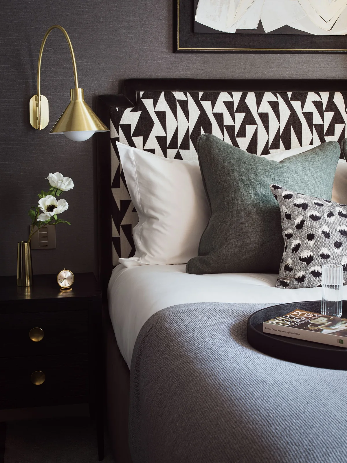 A black-and-white angular patterned headboard sits against a dark wall beside a gold-colored wall lamp and dark bedside drawers with pale and gray pillows