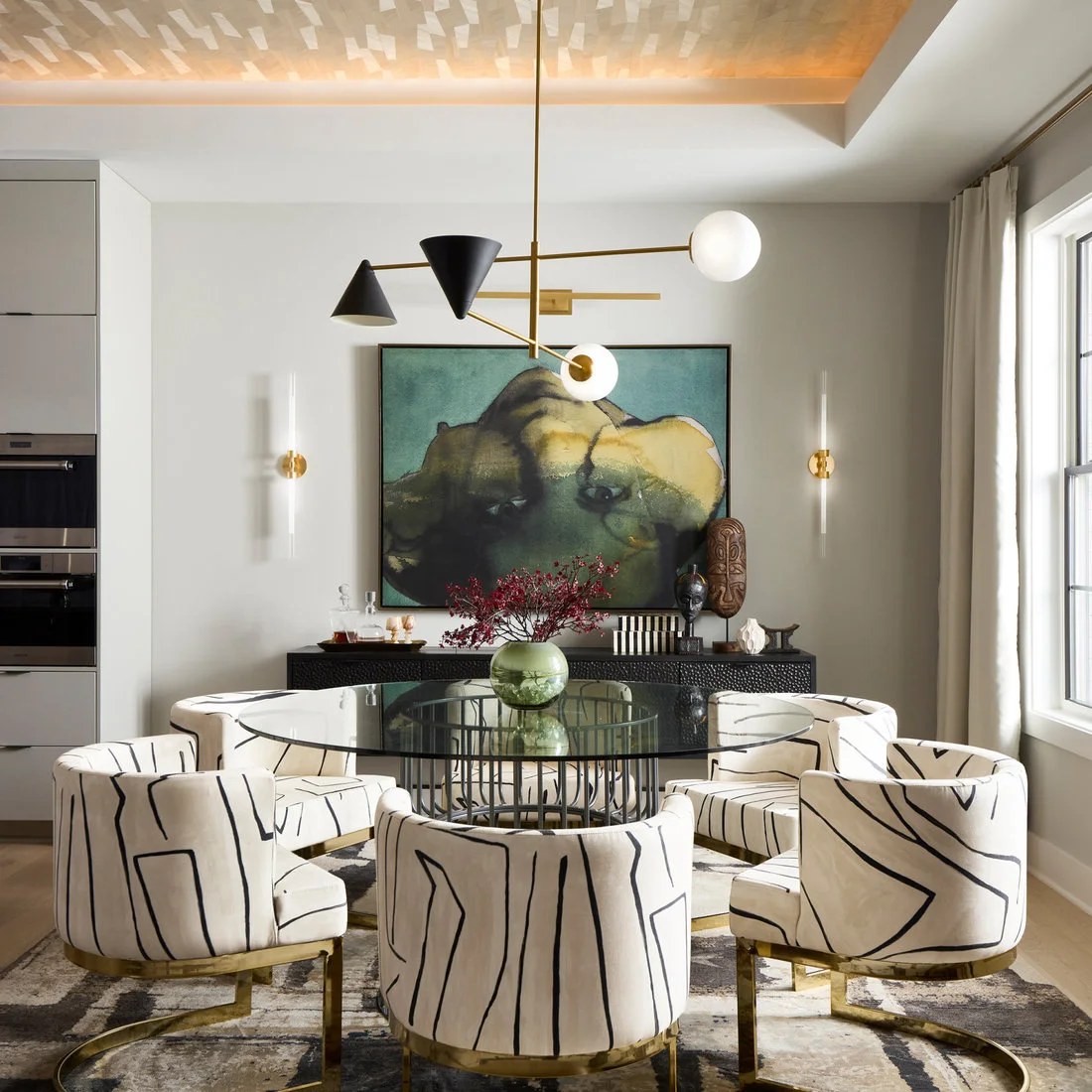 A dark sideboard sits beneath a large green-toned artwork behind a round glass dining table and pale chairs with black line patterns