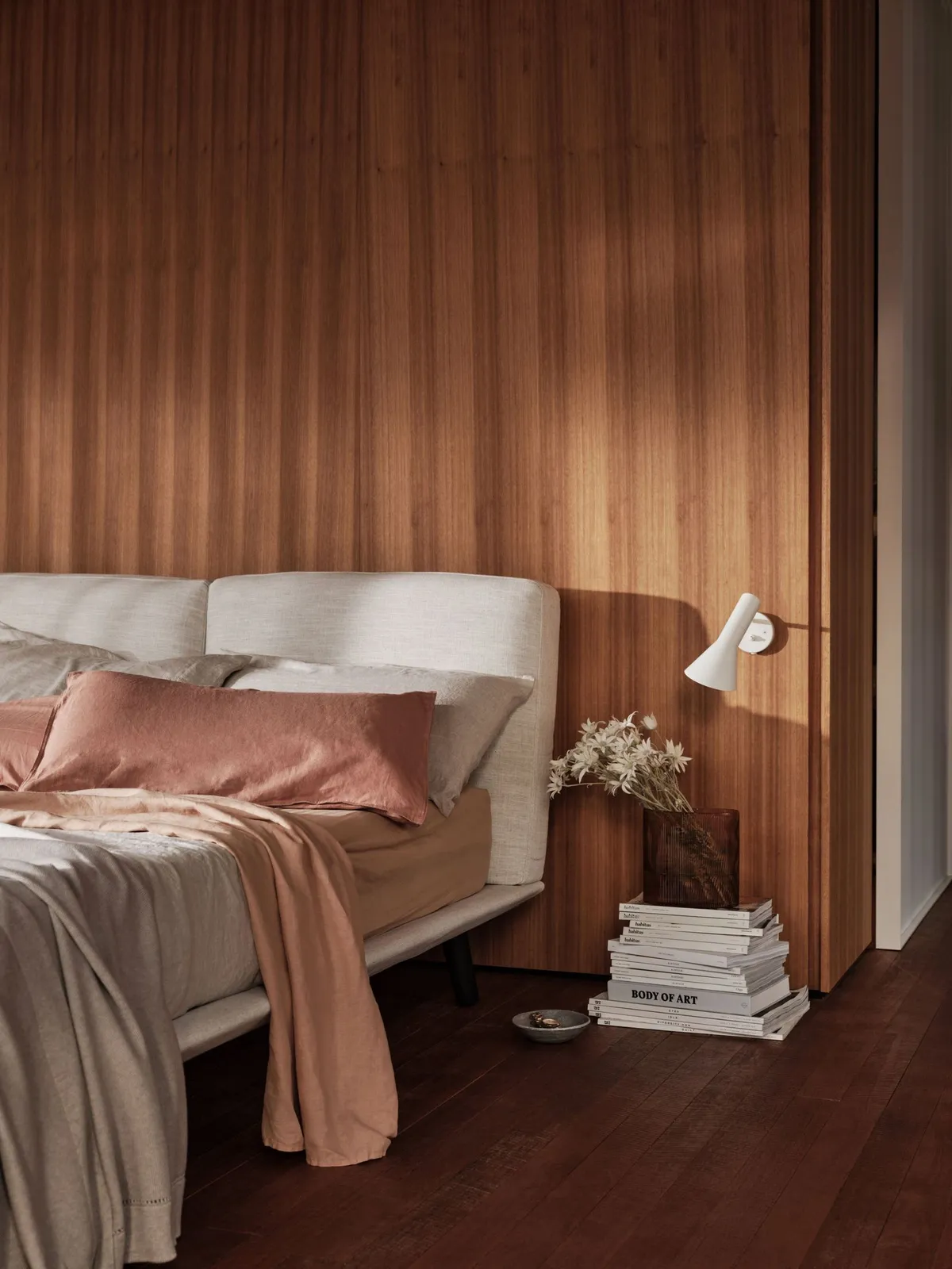 A pale bed with soft pink layers sits against vertical timber paneling.