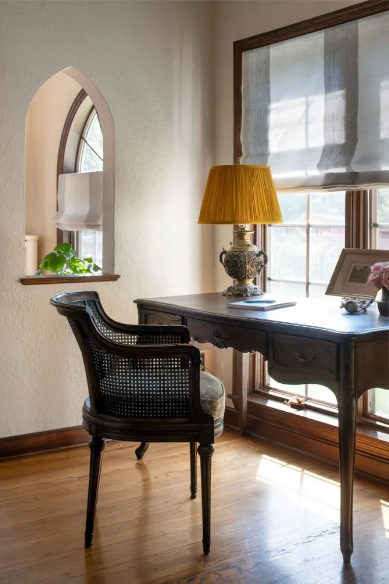Dark wood writing desk and woven-back chair beside window with shade, yellow-shaded lamp and arched interior opening