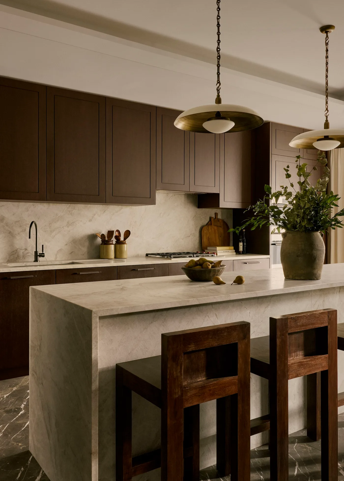 Brown paneled kitchen cabinets behind a pale waterfall-style island, wooden stools, broad metal pendants, pale backsplash, and black tap.