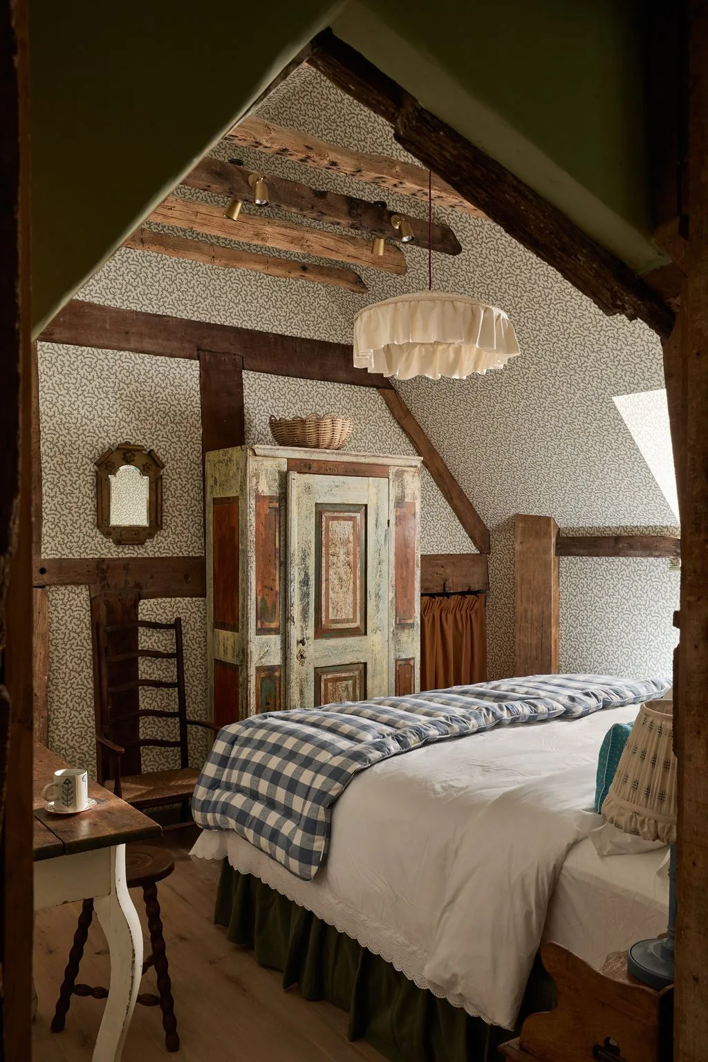 Cottage bedroom with exposed dark beams, small-pattern wallpaper, painted cupboard, blue checked blanket and pale ruffled pendant