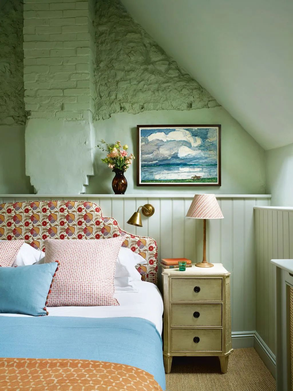 Bedroom with pale green walls and paneling, exposed rough masonry, floral headboard, blue bedding and narrow pale bedside drawers