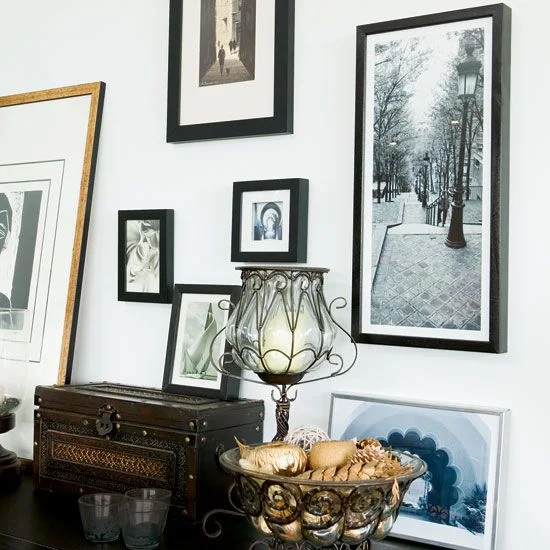 Black picture frames with pale mounts hang above a dark surface holding a decorative lamp, framed artwork and a warm-colored bowl beside a gold-colored mirror