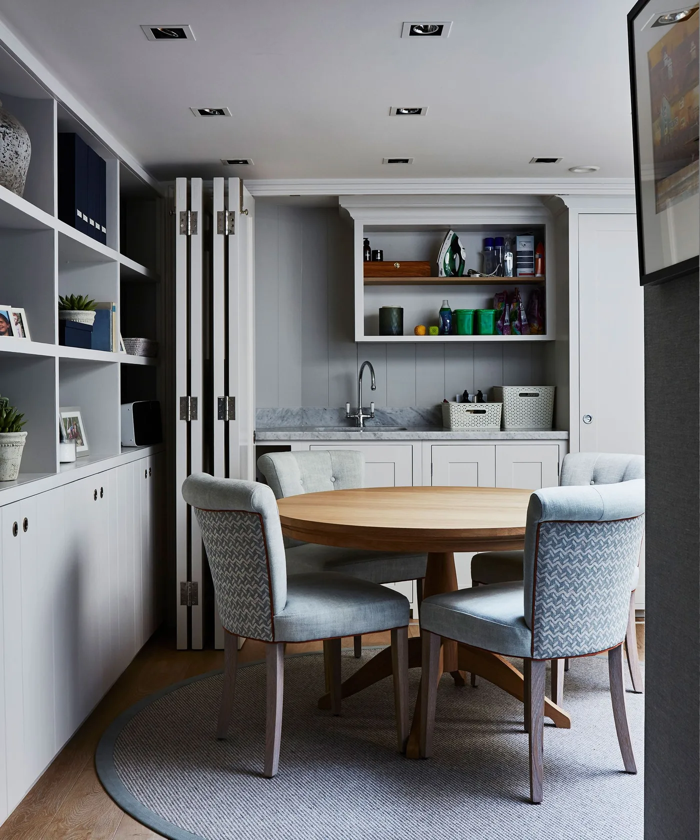 Pale dining cupboards and open shelves beside a round wood table with folding doors around a sink recess