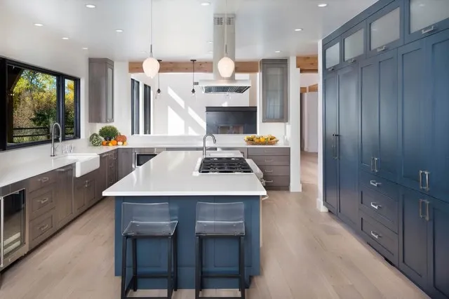 A full-height navy cabinet wall stands beside a navy island with pale counter and two stools facing wood-toned perimeter storage and pendant lights