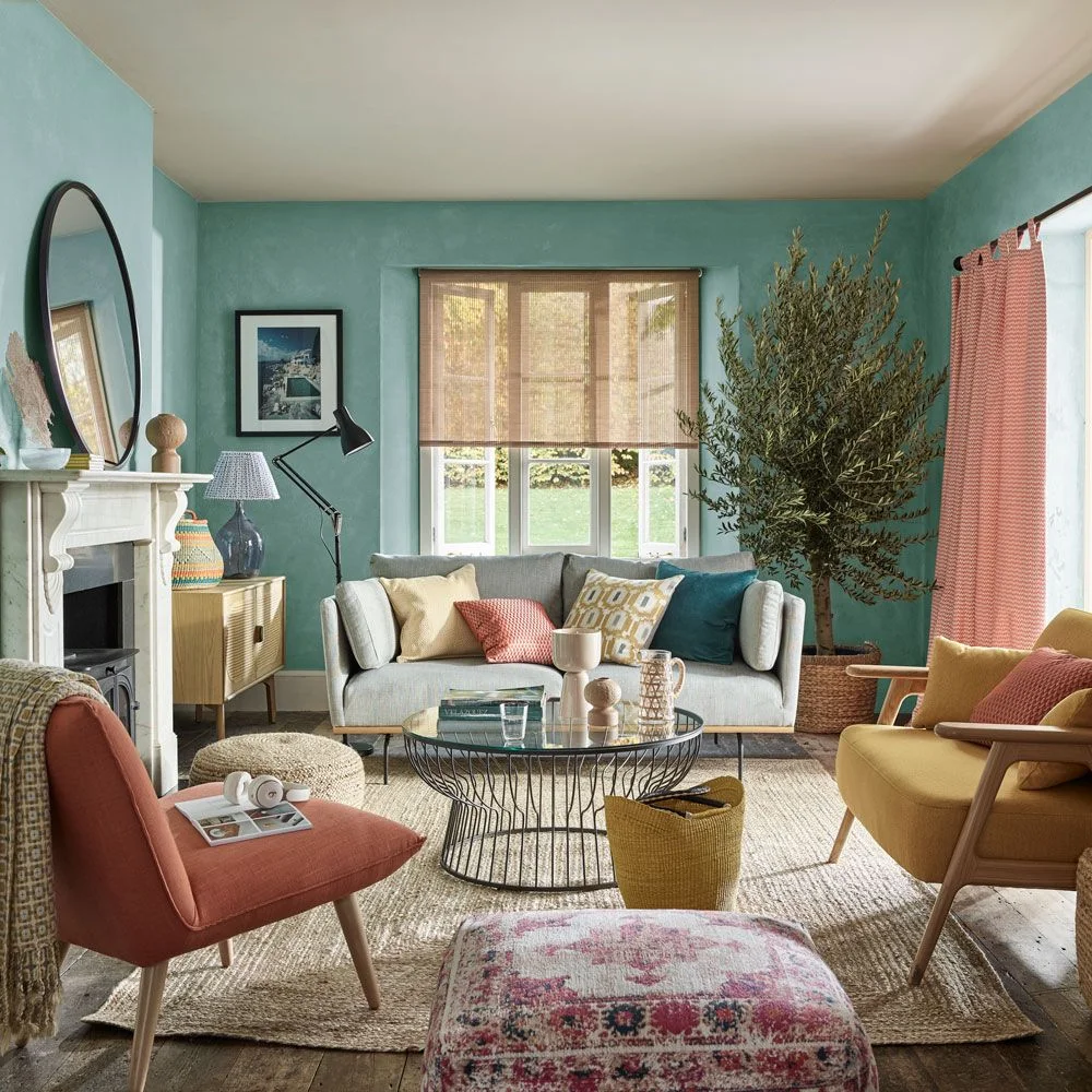 A pale gray sofa stands against teal walls beside pink and yellow chairs, a round glass-topped table and a white fireplace