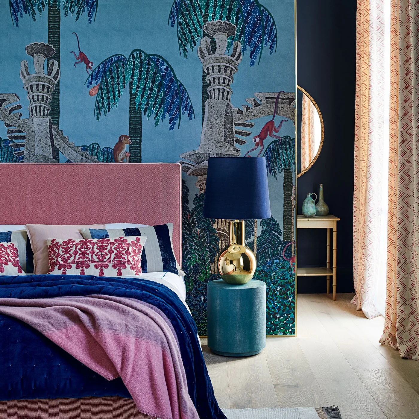A blue tropical wallpaper scene with palms and architectural shapes sits behind a pink headboard, blue bedding and a blue-shaded gold-colored lamp