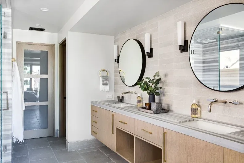 Light wood double vanity beneath a pale tiled wall with round dark-framed mirrors and a striped counter